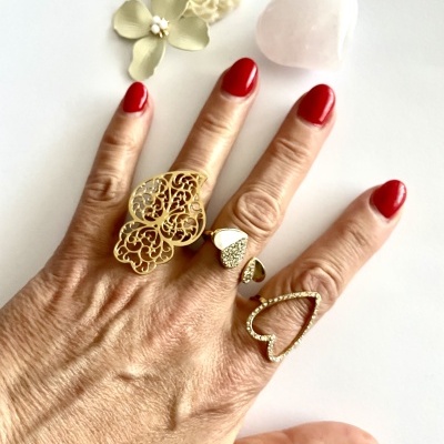 Mão com três anéis dourados, unhas vermelhas, flores branca e bege, coração de cristal rosa em fundo branco