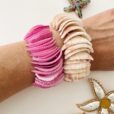 Dois braceletes de conchas, um rosa e outro creme, usados nos pulsos, com decorações marinhas e florais ao fundo.