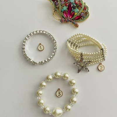 Quatro peças de joalharia sobre fundo branco, incluindo broche colorido e pulseiras de pérolas e prateadas numeradas.