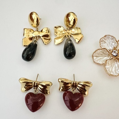 Dois pares de brincos dourados com laço, pedra preta em gota e pedra vermelha em coração, sobre fundo branco com flor decorativa.