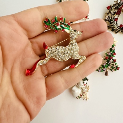 Broche de rena brilhante com detalhes coloridos numa mão sobre fundo branco com outros broches de Natal.