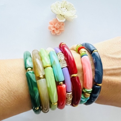 Pulseiras coloridas tubulares com detalhes dourados num pulso, fundo branco com duas flores decorativas