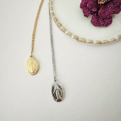 Duas correntes com pendentes ovais religiosos, dourada e prateada, sobre fundo branco com decoração bordada e flor roxa