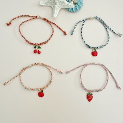 Quatro pulseiras trançadas com pendentes de frutas sobre fundo claro com estrela-do-mar e flor azul
