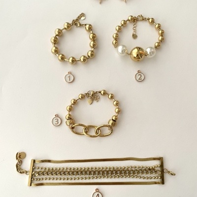 Cinco peças de bijuteria douradas numeradas, incluindo pulseiras e bracelete, com brincos de abelha ao fundo.