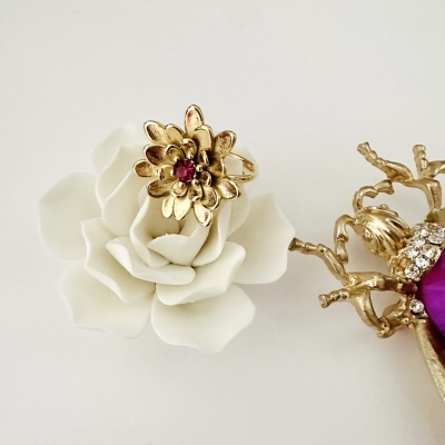 Anel dourado em forma de flor com pedra vermelha sobre flor branca decorativa próximo a joia dourada com pedras