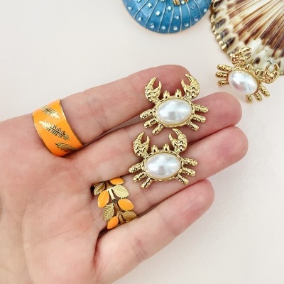 Brincos de caranguejo dourados com pérolas, mão com anéis alaranjados e conchas decorativas