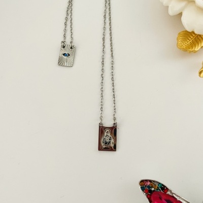 Dois colares com pendentes retangulares metálicos sobre fundo branco com flor decorativa e detalhe colorido