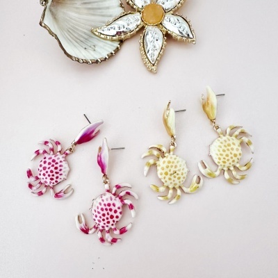 Brincos em forma de caranguejo rosa e amarelo sobre fundo branco com concha e flor decorativa