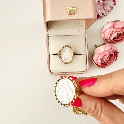 Anel dourado com figura religiosa em pedra branca com caixa cor de rosa e flores ao redor.