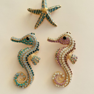 Três broches: uma estrela do mar azul turquesa e dois cavalos-marinhos com pedras azuis e rosas, fundo bege.