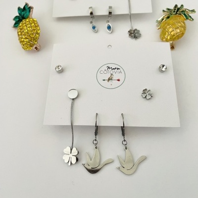 Brincos em metal com abacaxi amarelo, trevo de quatro folhas e pássaros prateados em cartões brancos com logo
