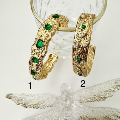 Dois braceletes dourados com pedras verdes e cristais sobre uma taça de vidro, com uma libélula decorativa translúcida e glitter prateado à frente