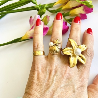 Mão com três anéis dourados decorativos, unhas vermelhas e flores ao fundo