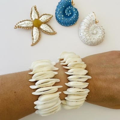Pulseiras de conchas brancas naturais no pulso e três broches decorativos em fundo branco.