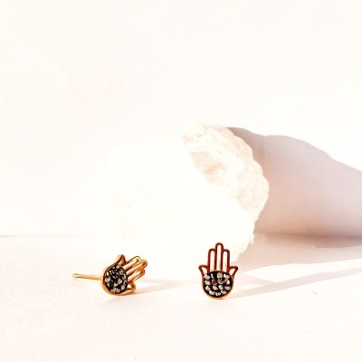Brincos dourados em forma de mão com pedras sobre superfície branca.