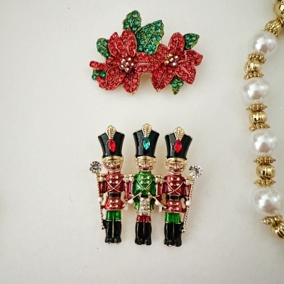 Broches decorativos de soldadinhos e flores com pedras coloridas, parcialmente ao lado pérolas brancas e douradas.