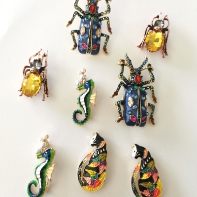 Broches coloridos em forma de insetos, cavalos-marinhos e gatos, com pedras e detalhes metálicos, sobre fundo branco