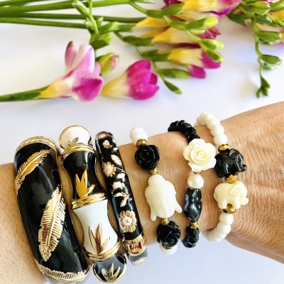 Pulseiras em metal dourado e contas pretas e brancas com formas de tartaruga, flor e elefantes no pulso, com flores ao fundo