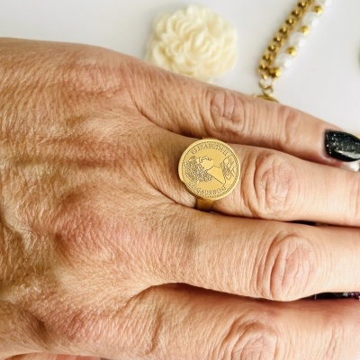 Anel dourado redondo com figura e texto, usado numa mão com acessórios ao fundo