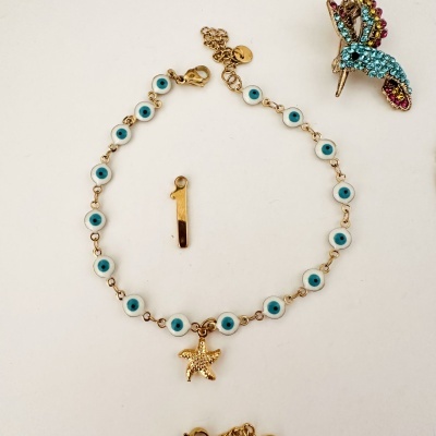 Pulseira com elementos de olhos azuis e pendente de estrela do mar dourada com um brinco de beija-flor colorido em fundo branco