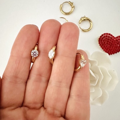 Três brincos dourados com pedras brilhantes segurados por uma mão e outros acessórios ao fundo