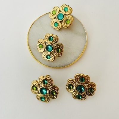 Conjunto dourado com pedras verdes e azuis em forma de flor sobre fundo branco com prato redondo dourado
