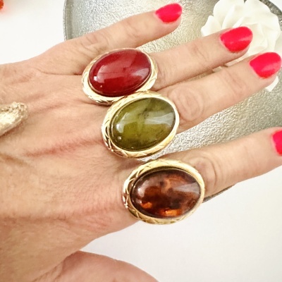 Anel com gemas grandes em vermelho, verde e castanho com borda dourada, em mão com unhas pintadas de rosa.