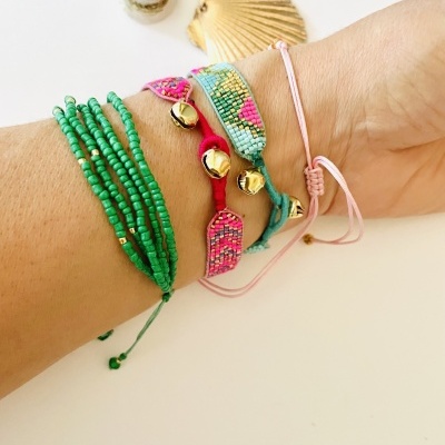 Pulseira verde de contas, duas pulseiras coloridas com sinos dourados, pulseira rosa de cordão fino num pulso com conchas ao fundo