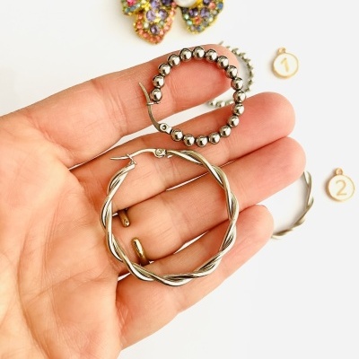 Brincos de argola em metal prateado, um com contas e outro torcido, sobre fundo branco com discos numerados e peça decorativa colorida.