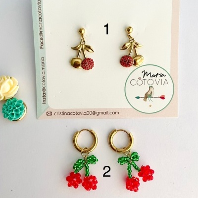 Dois pares de brincos em tema de cerejas, dourado e vermelho, com cartão de marca e informação de contacto.