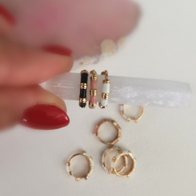 Brincos dourados com esmaltados preto, branco e rosa, dispostos sobre superfície branca com mão segurando uma peça e pedra rosa ao fundo