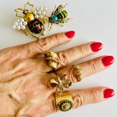 Mão com anéis dourados e broches de abelhas coloridas sobre fundo branco.