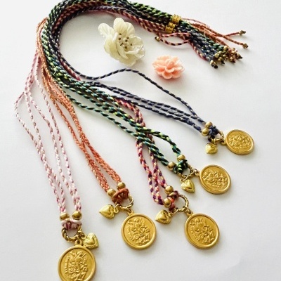 Seis colares com fios trançados coloridos e pendentes dourados em fundo branco com duas flores decorativas