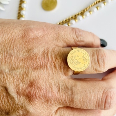 Anel dourado com placa redonda gravada em mão com unhas pretas e pulseiras douradas e pérolas ao fundo