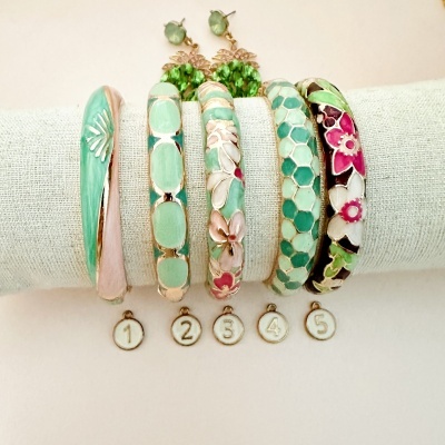 Cinco pulseiras esmaltadas coloridas expostas sobre suporte branco com etiquetas numeradas