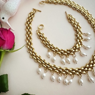 Conjunto de joias douradas com correntes e pérolas penduradas perante fundo claro com rosa e pássaro decorativo.