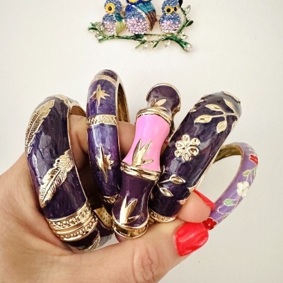 Pulseiras esmaltadas roxas com detalhes dourados e rosa, mão segurando, broche de corujas ao fundo