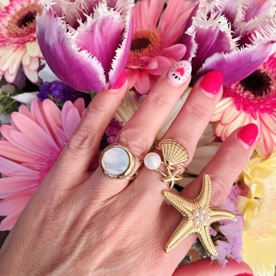 Mão feminina com anéis dourados e unhas decoradas segurando tulipa roxa com flores cor-de-rosa ao fundo.