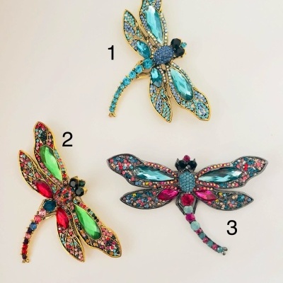 Três broches coloridos em forma de libélulas com pedras brilhantes em azul, verde, vermelho e rosa.