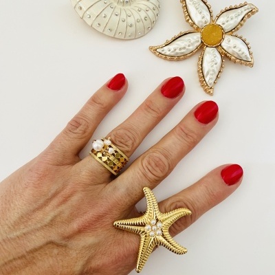 Mão com unhas vermelhas a usar anéis dourados com pérola e forma de estrela do mar, com broches dourados em concha e flor numa superfície branca