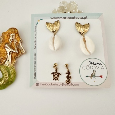 Conjunto de brincos com conchas e pendentes dourados, em cartão de exibição, ao lado de figura de sereia.