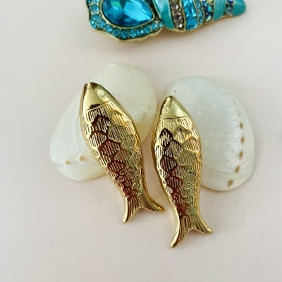 Brincos dourados em forma de peixe com conchas brancas ao fundo