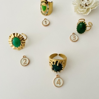 Quatro anéis dourados com pedras verdes e etiquetas numeradas sobre fundo branco com flor decorativa