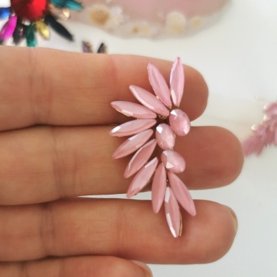 Brinco rosa em forma de pétalas brilhantes com acabamento dourado segurado pelos dedos com outros brincos coloridos ao fundo