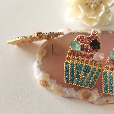 Brincos dourados em forma de cupcake com pedras coloridas sobre superfície branca e base de pedra rosa