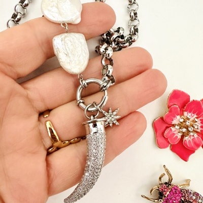 Colar metálico com pérolas e pendentes brilhantes numa mão ao lado de broches decorativos