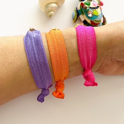 Três pulseiras elásticas coloridas em tecido no pulso da mão com ornamento colorido e concha ao lado.