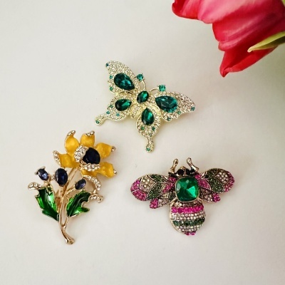 Três broches decorativos coloridos em fundo branco com flor vermelha no canto.