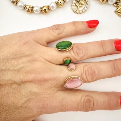Mão com anéis dourados e pedras verdes e rosa, unhas vermelhas, colar de pérolas e dourado ao fundo
